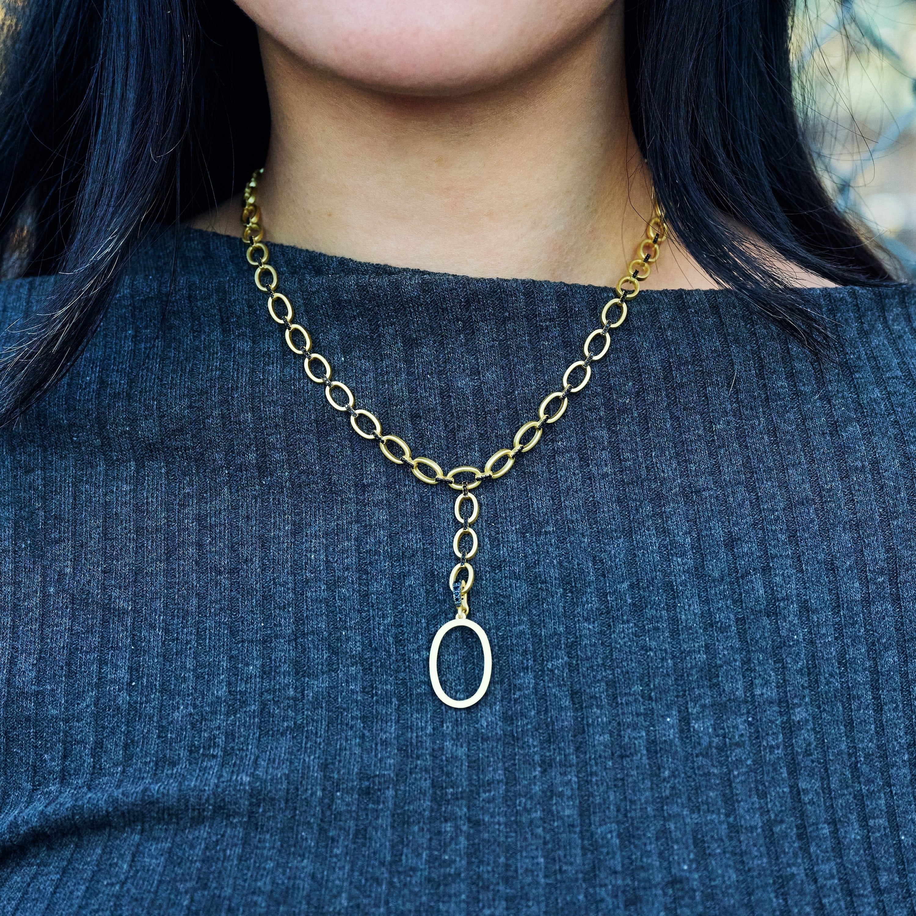 A person with long dark hair is wearing a ribbed dark gray top and the Industrial Finish Cobblestone Flat Chain Link Y-Necklace. The necklace, adorned with an oval pendant, hangs gracefully from the center of the distinctive cobblestone chain. The background is blurred, drawing focus to the beautiful necklace and elegant top.