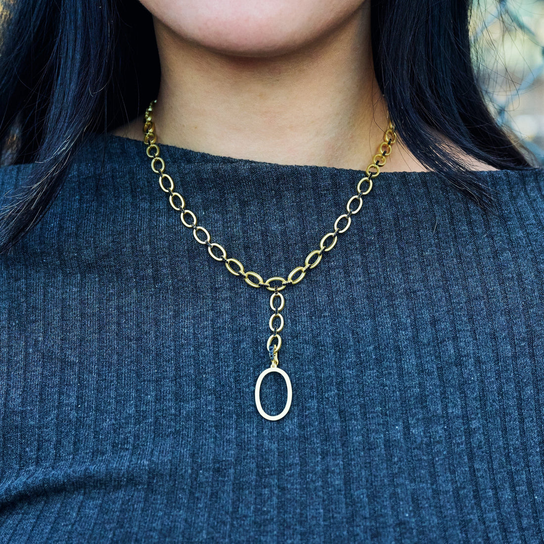 A person with long dark hair is wearing a ribbed dark gray top and the Industrial Finish Cobblestone Flat Chain Link Y-Necklace. The necklace, adorned with an oval pendant, hangs gracefully from the center of the distinctive cobblestone chain. The background is blurred, drawing focus to the beautiful necklace and elegant top.